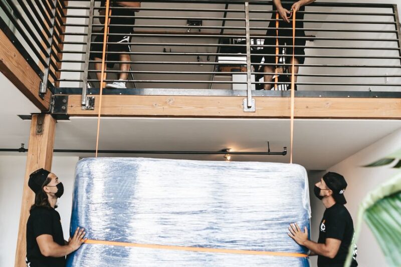 Two professional movers carry a wrapped sofa into a building stairwell.